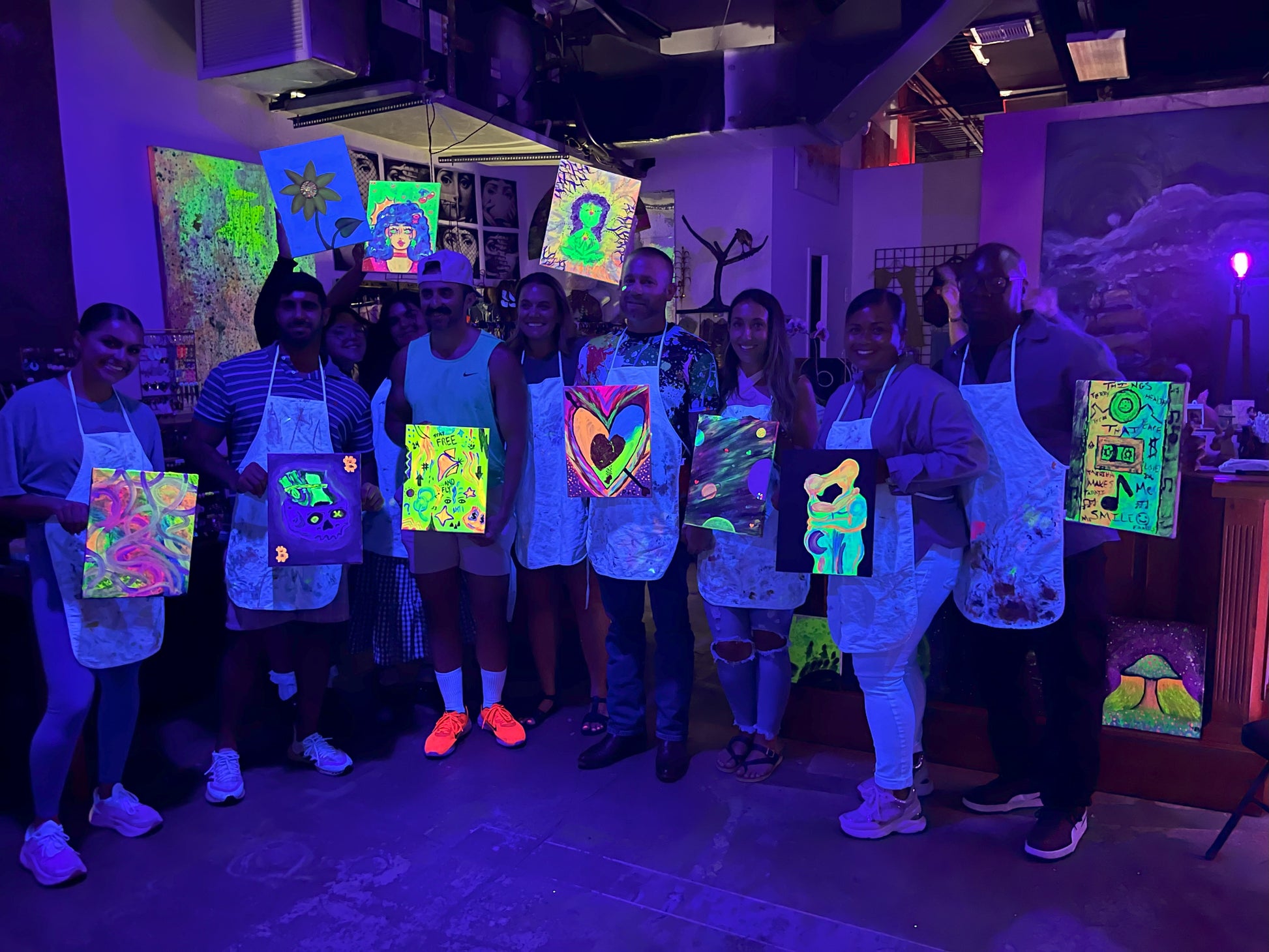 Group of guests proudly holding their neon paintings after a BYOB Neon Nights paint and sip session in Tampa.