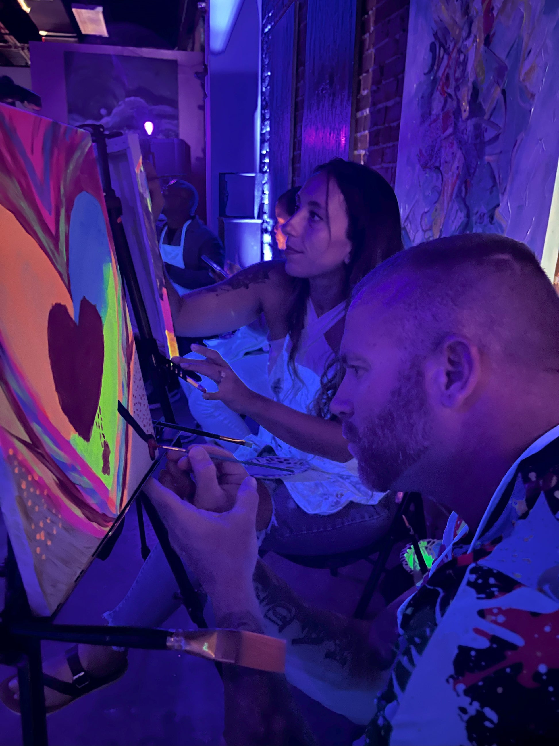 Guests painting glowing canvases under blacklights at Neon Nights BYOB sip and paint event at Marcolina's art gallery in Tampa.